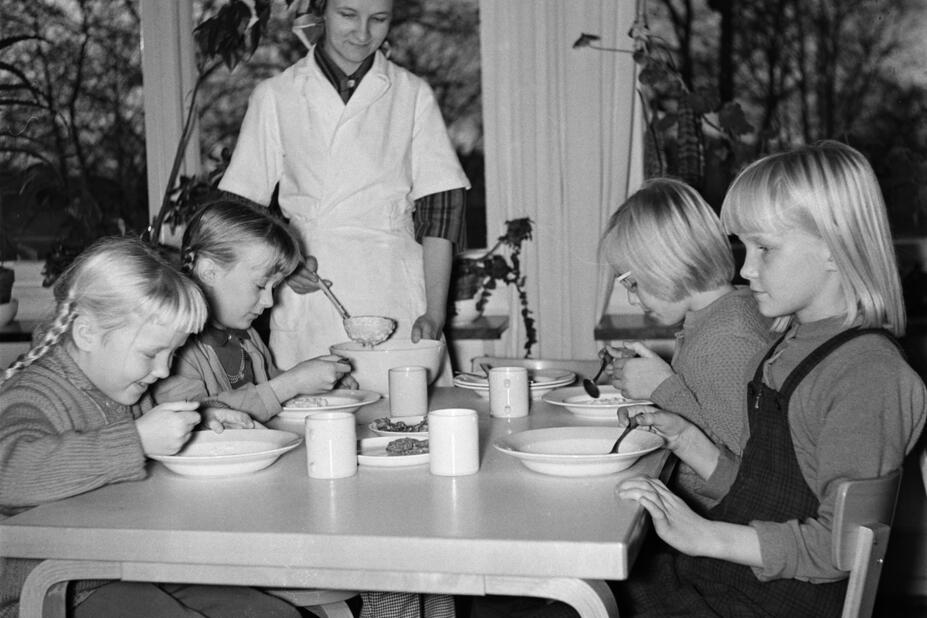 School children eating at the dining table with the food service worker watching next to them.