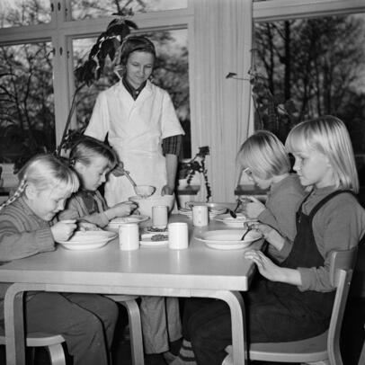 Koululaiset ruokapöydän äärellä ruokailemassa keittäjän katsoessa vieressä.